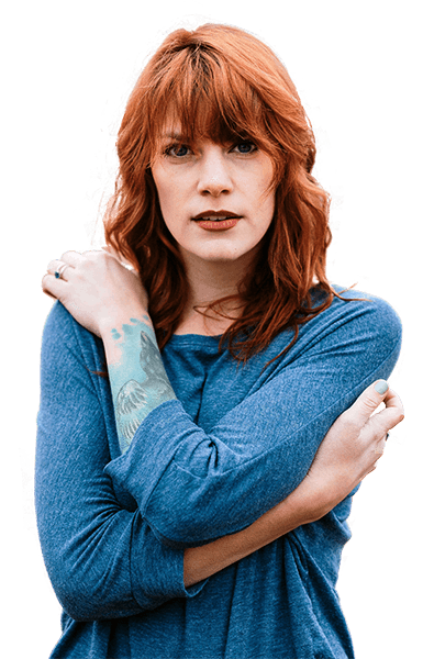 Red-haired woman with tattooed arm poses with crossed arms.